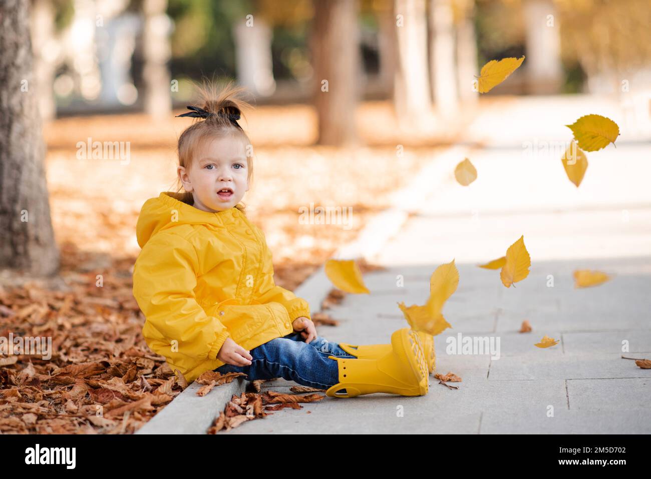 Cute funny kid girl 2-3 year old wear yellow rain jacket and rubber boots in autumn park outdoor over fallen leaves. Fall season. Childhood. Stock Photo