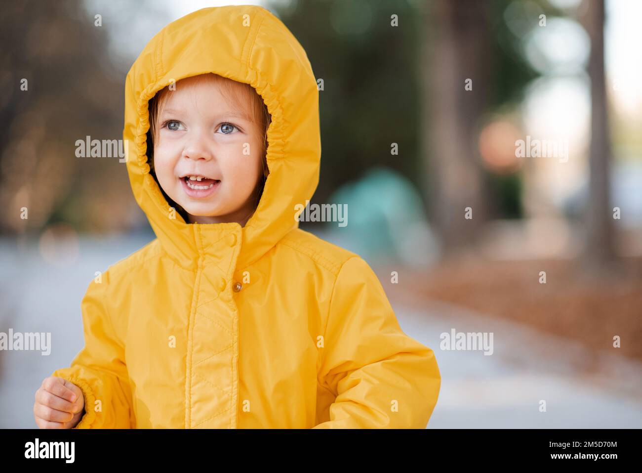 Cute funny kid girl 2-3 year old wear yellow rain jacket with hood in autumn park outdoor. Fall season. Childhood. Stock Photo