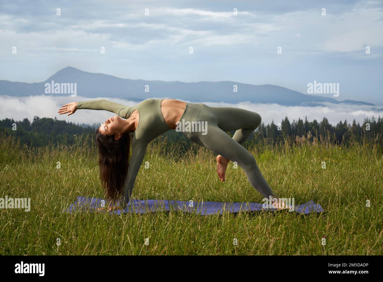Active young woman in sport clothes spending free time for workout on fresh air. Dark haired caucasian lady doing flexible exercises on yoga mat. Stock Photo