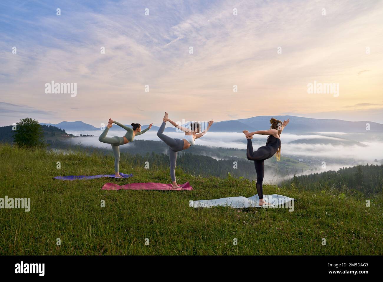 Three sporty women doing exercises for balance on yoga mat amonu summer mountains. Group of young females in sport clothes having outdoors workout together. Stock Photo