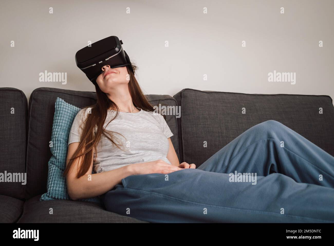 Beautiful smiling young woman wearing VR goggles and virtually traveling through the metaverse from the comfort of her couch Stock Photo