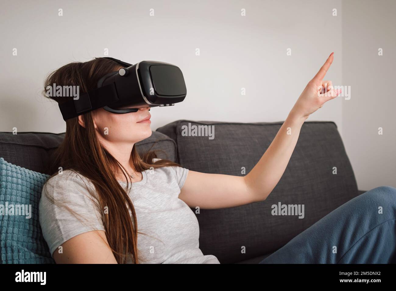 Beautiful smiling young woman wearing VR goggles and virtually traveling through the metaverse from the comfort of her couch Stock Photo
