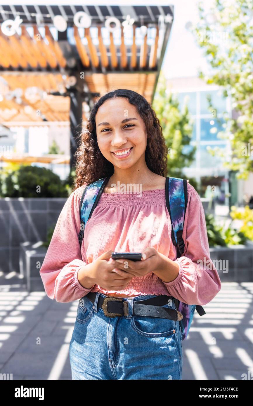 Latin university student woman smiling and using her mobile sitting outside the campus in free