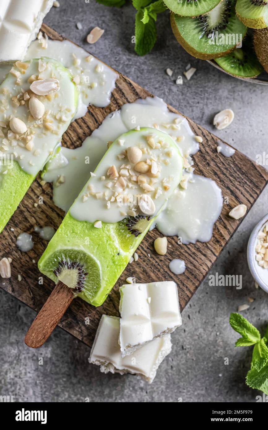 Homemade vegan kiwi ice cream bars or popsicles covered with white chocolate and nuts on wooden board, top view, flat lay. Vertical Stock Photo