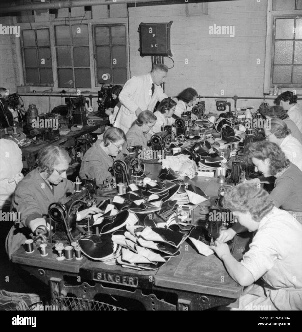 At a factory somewhere in the Midlands, machinists sew together the component parts of the outsides of the shoes they are manufacturing for members of the Women's Royal Naval Service.  In the background, the overseer can be seen, inspecting the work of one of the women. Stock Photo