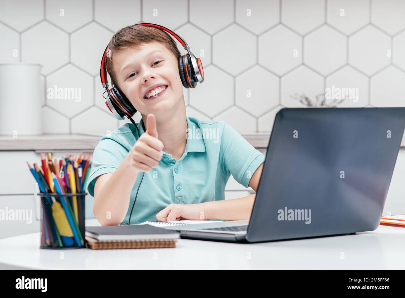 Portrait of young preteen boy wearing blue T-shirt, black red headphones, sitting at desk in front of laptop near houseplant, showing thumb up sign Stock Photo