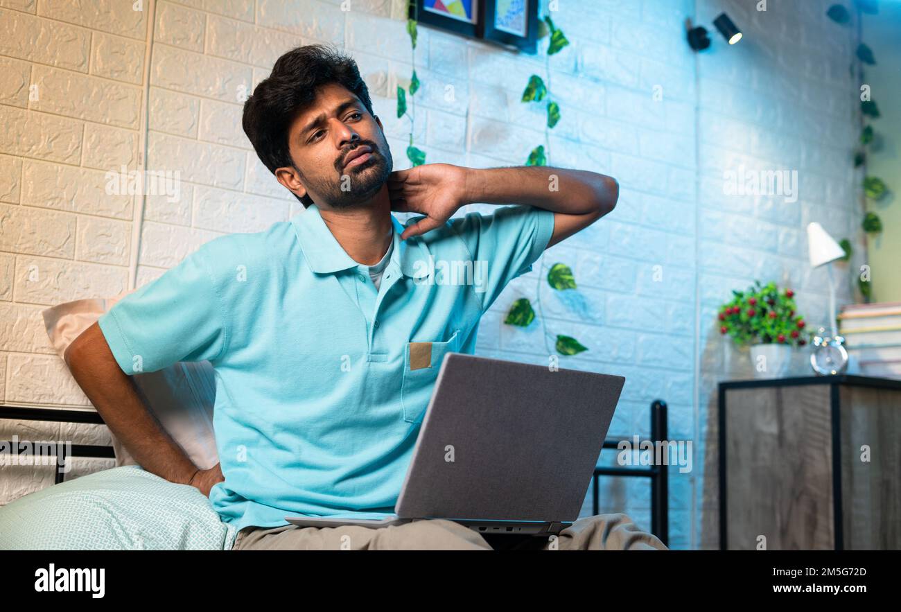 young man suffering from neck and back pain while using laptop on bed during work from home at night - concept of improper working posture, overworked Stock Photo