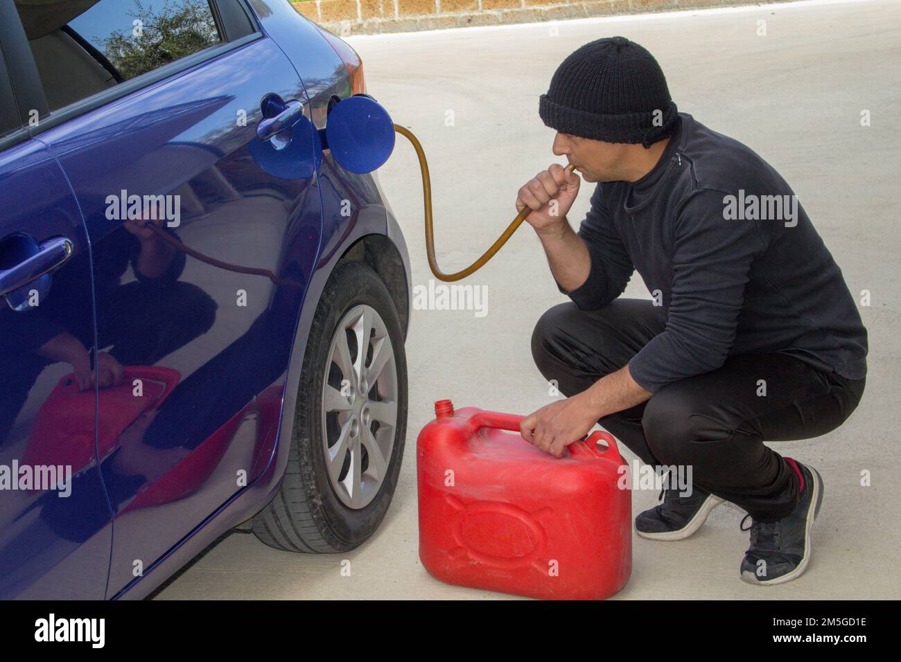 Image of a thief dressed in black who sucks petrol from a car tank with