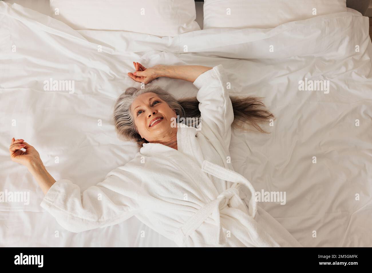 Senior woman in a bathrobe looking up while lying on a bed. Smiling retired female enjoying morning. Stock Photo
