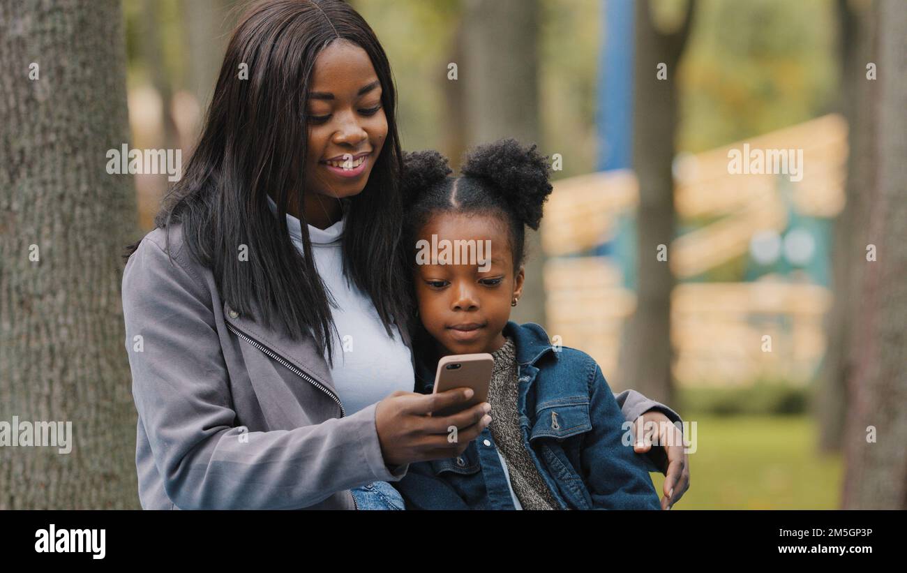 African American woman mother with child daughter little kid girl small baby schoolgirl sitting in park outdoors browsing mobile phone scrolling Stock Photo