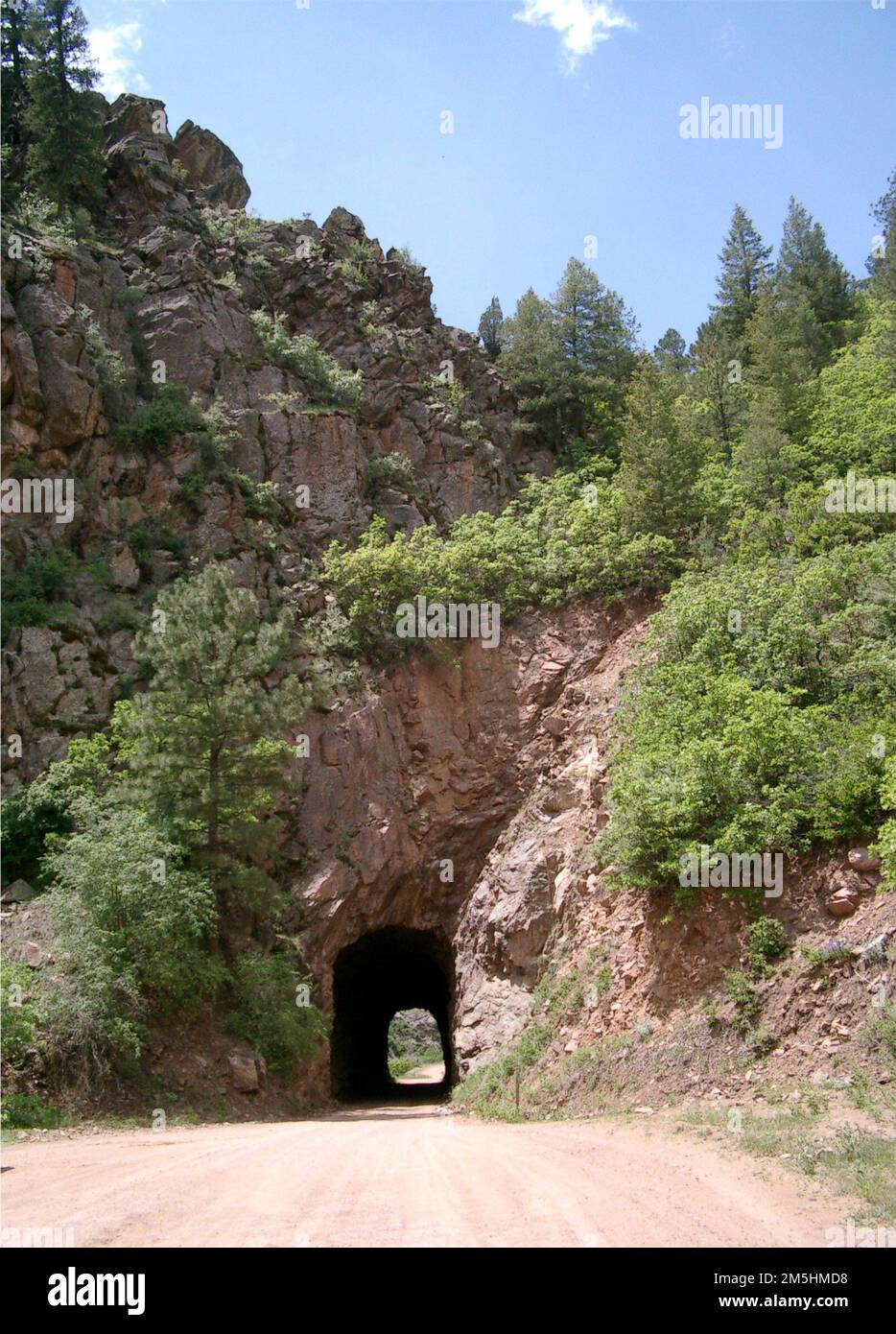 . Phantom Canyon Road, a dirt-surfaced road, passes through a narrow tunnel carved through a pale red-rock slope on a sunny day in summer. Colorado (38.520° N 105.125° W) Stock Photo