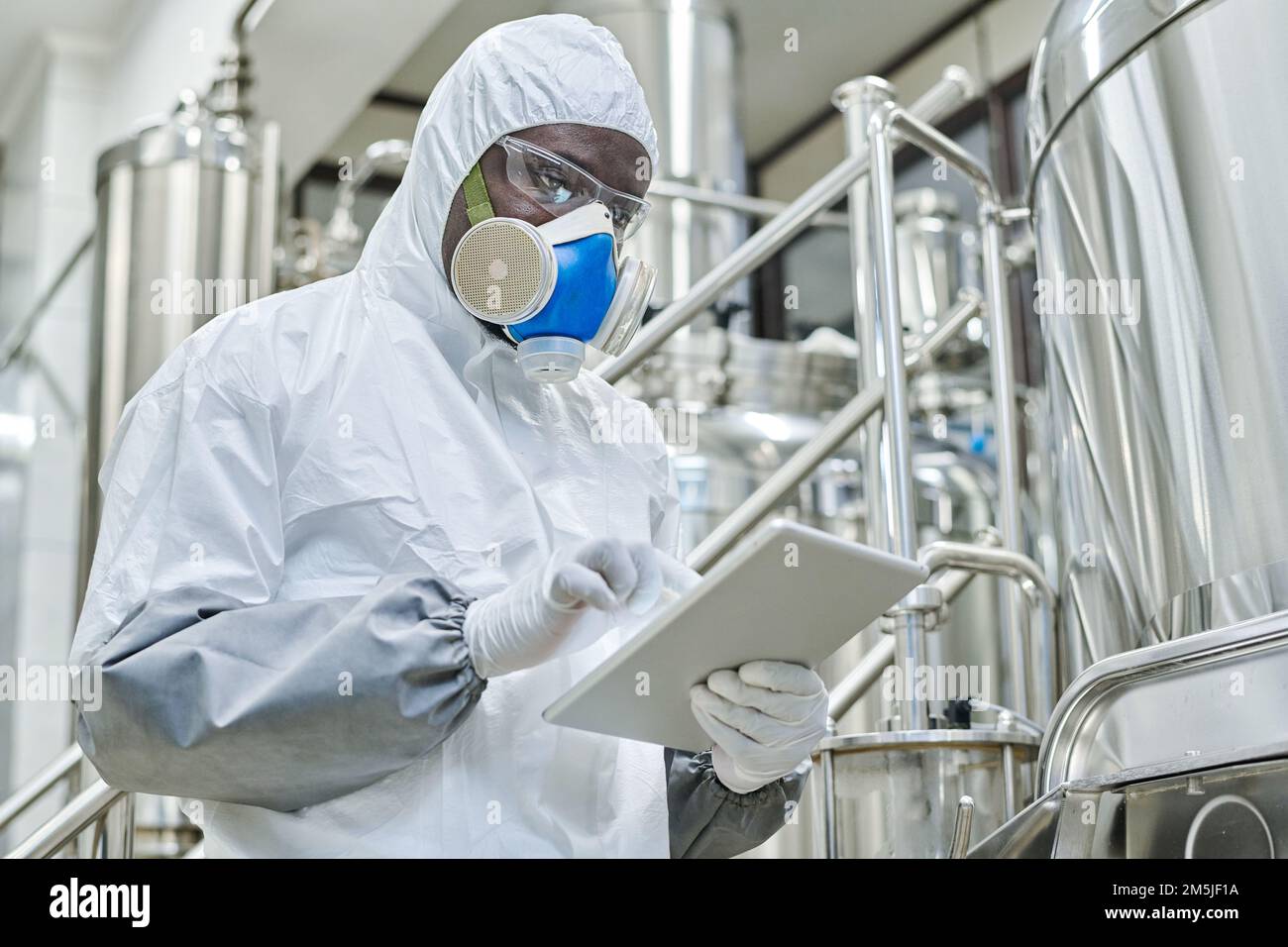 African American chemist in protective costume working online on tablet pc during his work on chemical production Stock Photo