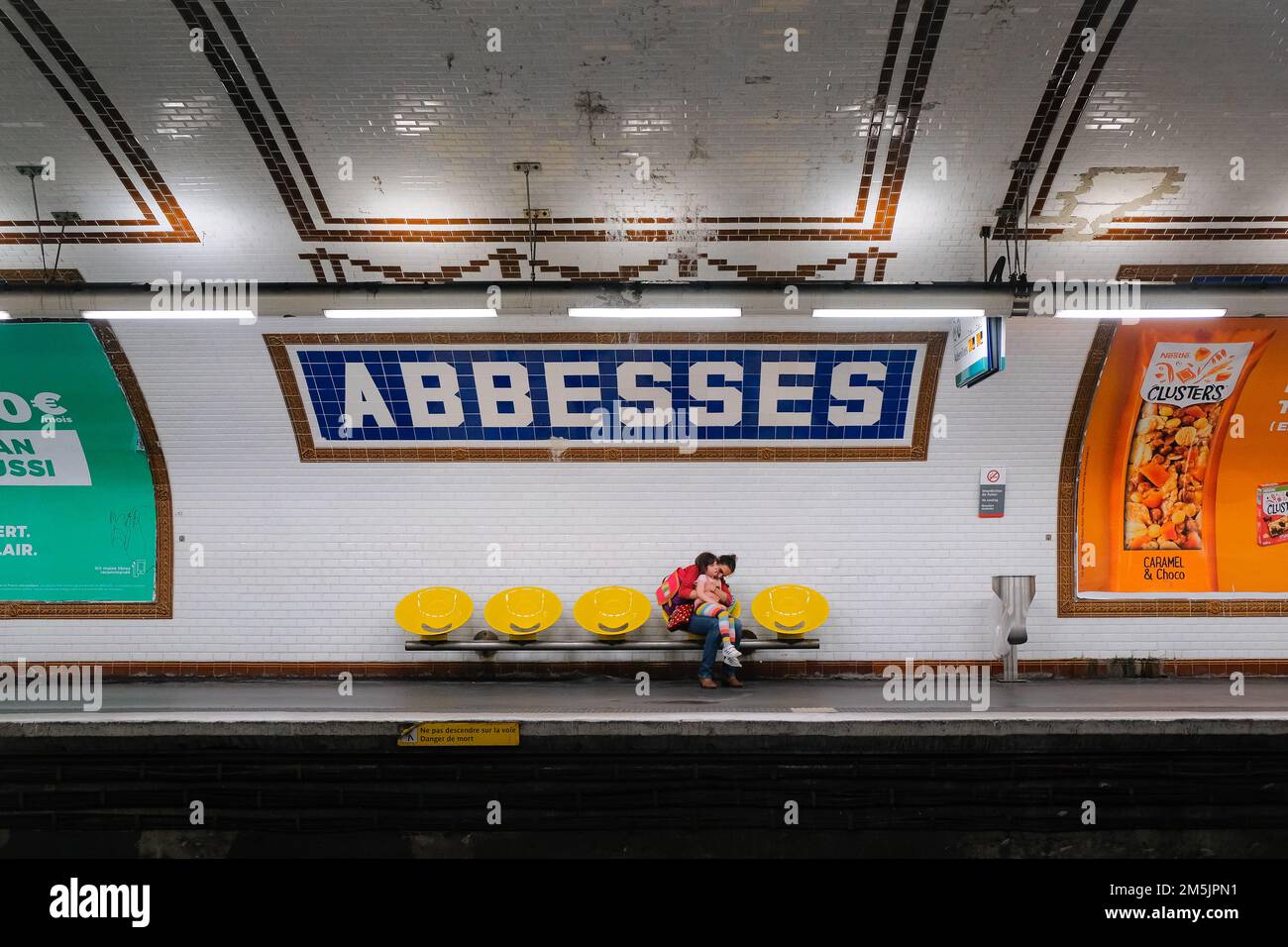 Paris, France - mother sits on yellow chair, cuddles daughter on lap. Both wait for next train inside Abbesses metro station platform in Montmartre. Stock Photo
