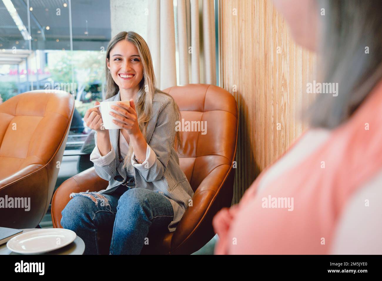 Happy young woman sitting on an armchair, talking and sharing coffee with a friend in open workplace Stock Photo