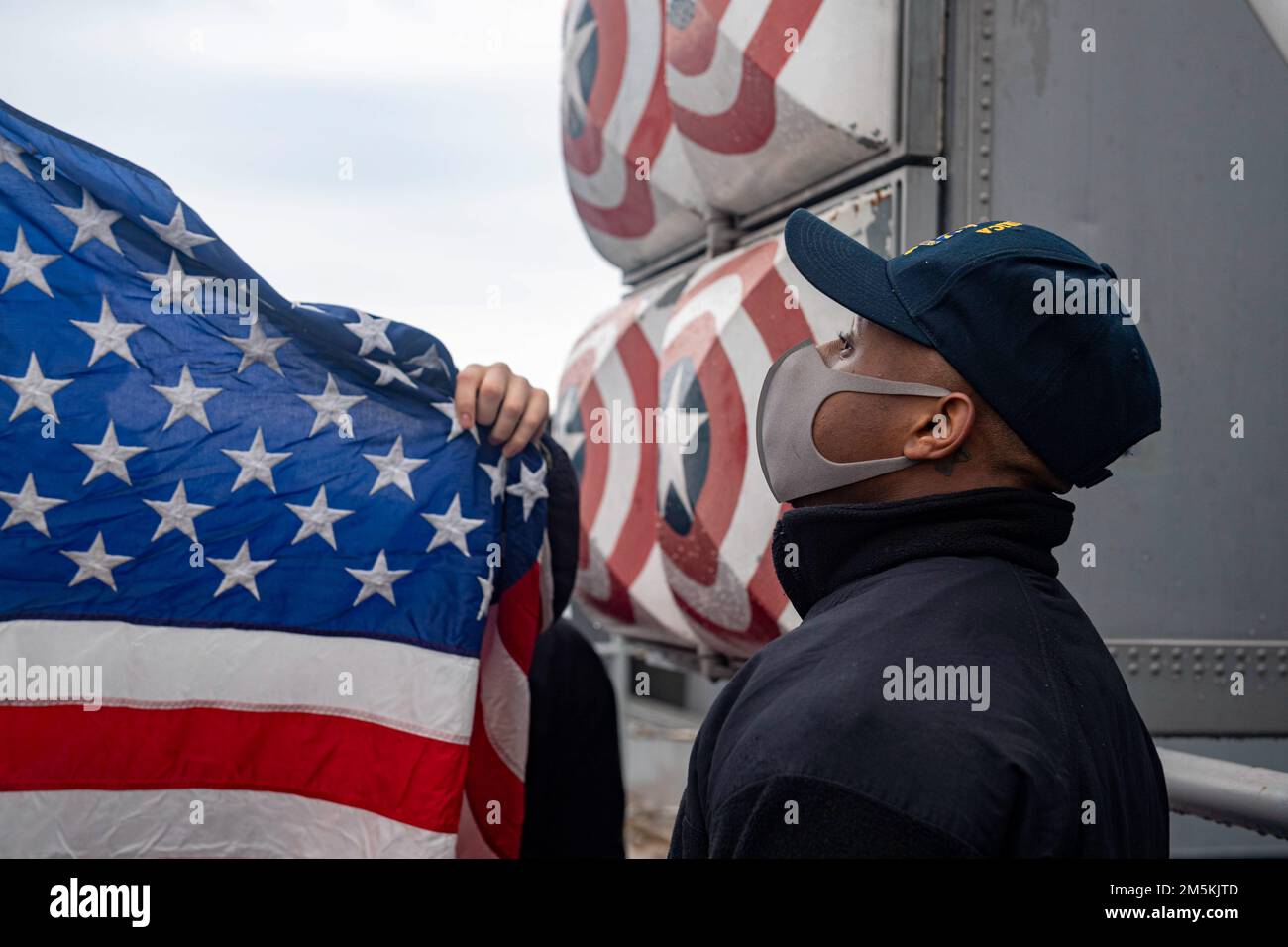 SASEBO, Japan (March 22, 2022) Aviation Boatswain’s Mate (Handling) Airman Camron Lamarre, from Brooklyn, N.Y., assigned to the forward-deployed amphibious assault ship USS America (LHA 6), watches as the American flag is prepared for colors. America, lead ship of the America Amphibious Ready Group, is operating in the U.S. 7th Fleet area of responsibility to enhance interoperability with allies and partners and serve as a ready response force to defend peace and stability in the Indo-Pacific region. Stock Photo
