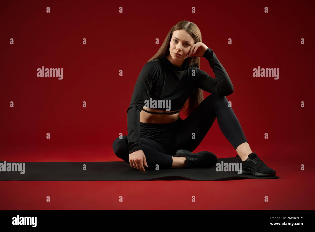 Front view of pretty brunette woman wearing in black sport suite sitting resting after training on yoga mat. Sporty fit female lean on knee looking at camera, relaxing on floor. Concept of sport. Stock Photo