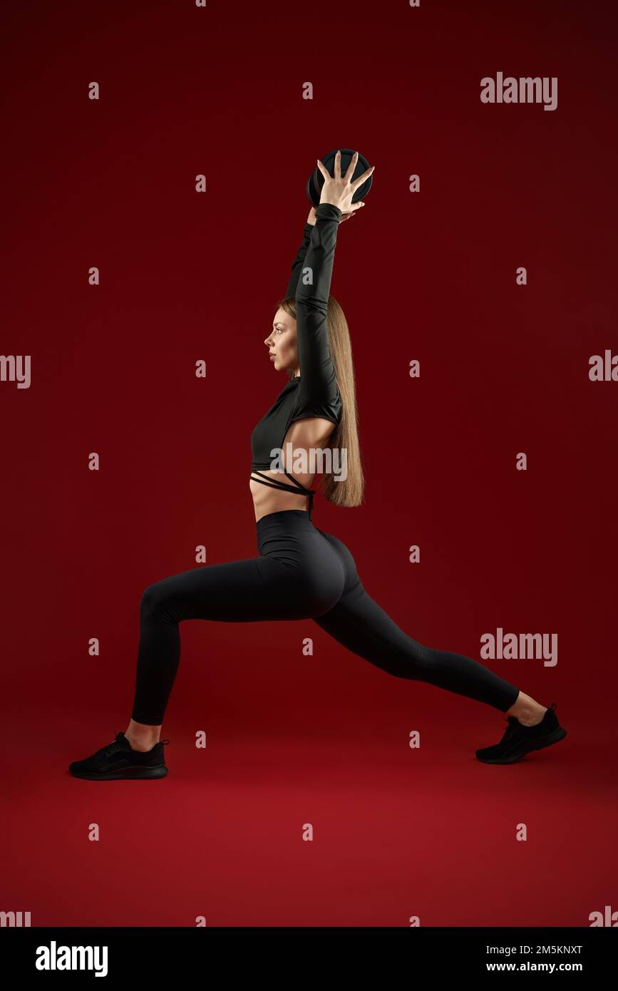 Side view of strong brunette woman in black suite doing training with weight dumbbell under head. Seductive and fit female training doing lunges in studio on red background. Stock Photo