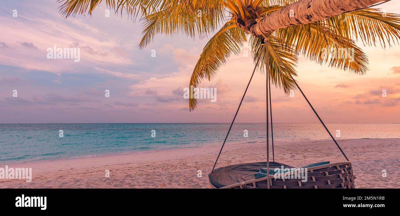Tropical sunset beach panorama as summer landscape with relax beach swing or hammock on coconut palm, white sand and calm sea beach tranquility banner Stock Photo
