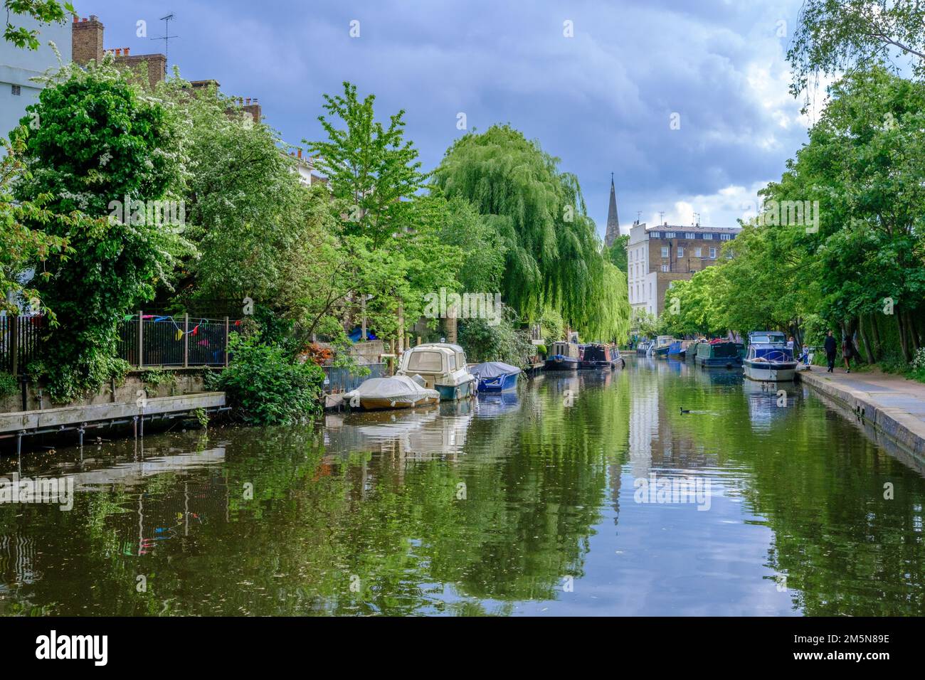 Boats docked on both sides of Regent’s Canal between Gloucester Ave & Regent’s Park Road, London. Spring foliage & trees give a serene atmosphere. Stock Photo