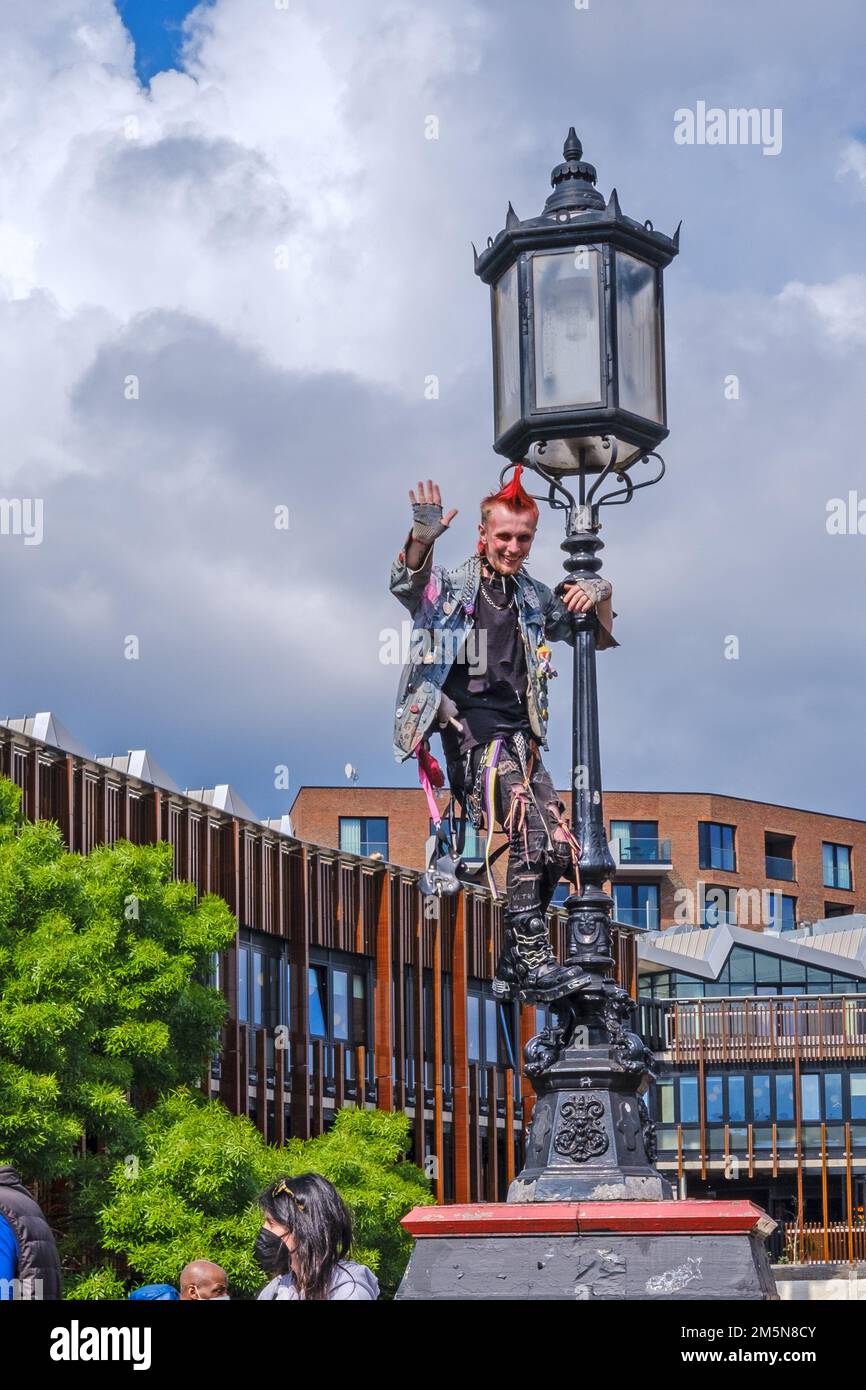 A man in punk style clothes & spiked mohawk hair smiles & waves from a lamppost where he has climbed in Camden Town, London. Stock Photo