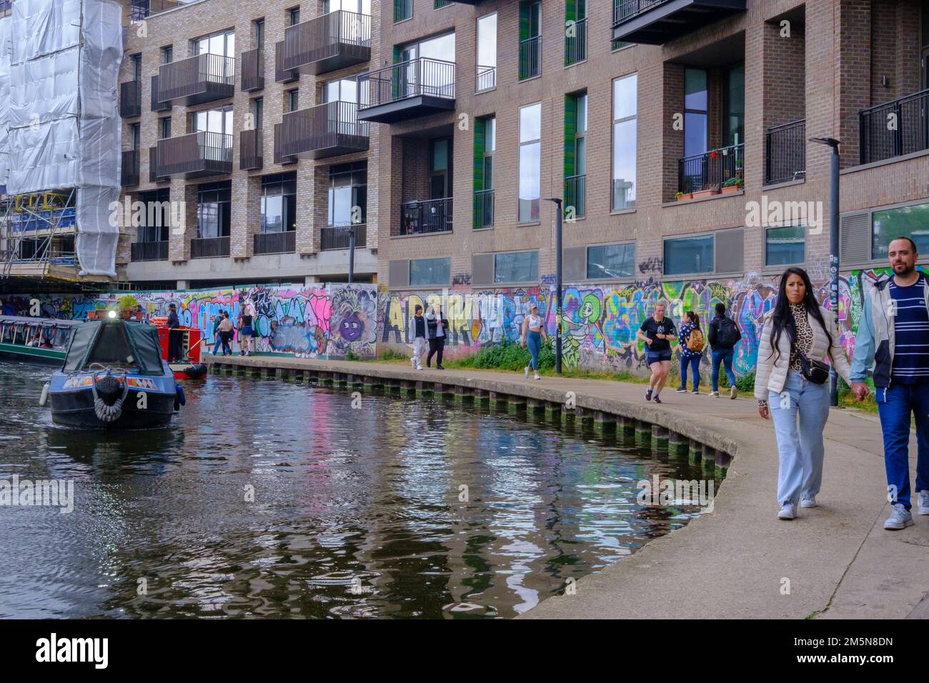 People walk & jog along Regents Canal towpath near Camden Town, with walls covered in graffiti and narrow boats in the water. Stock Photo