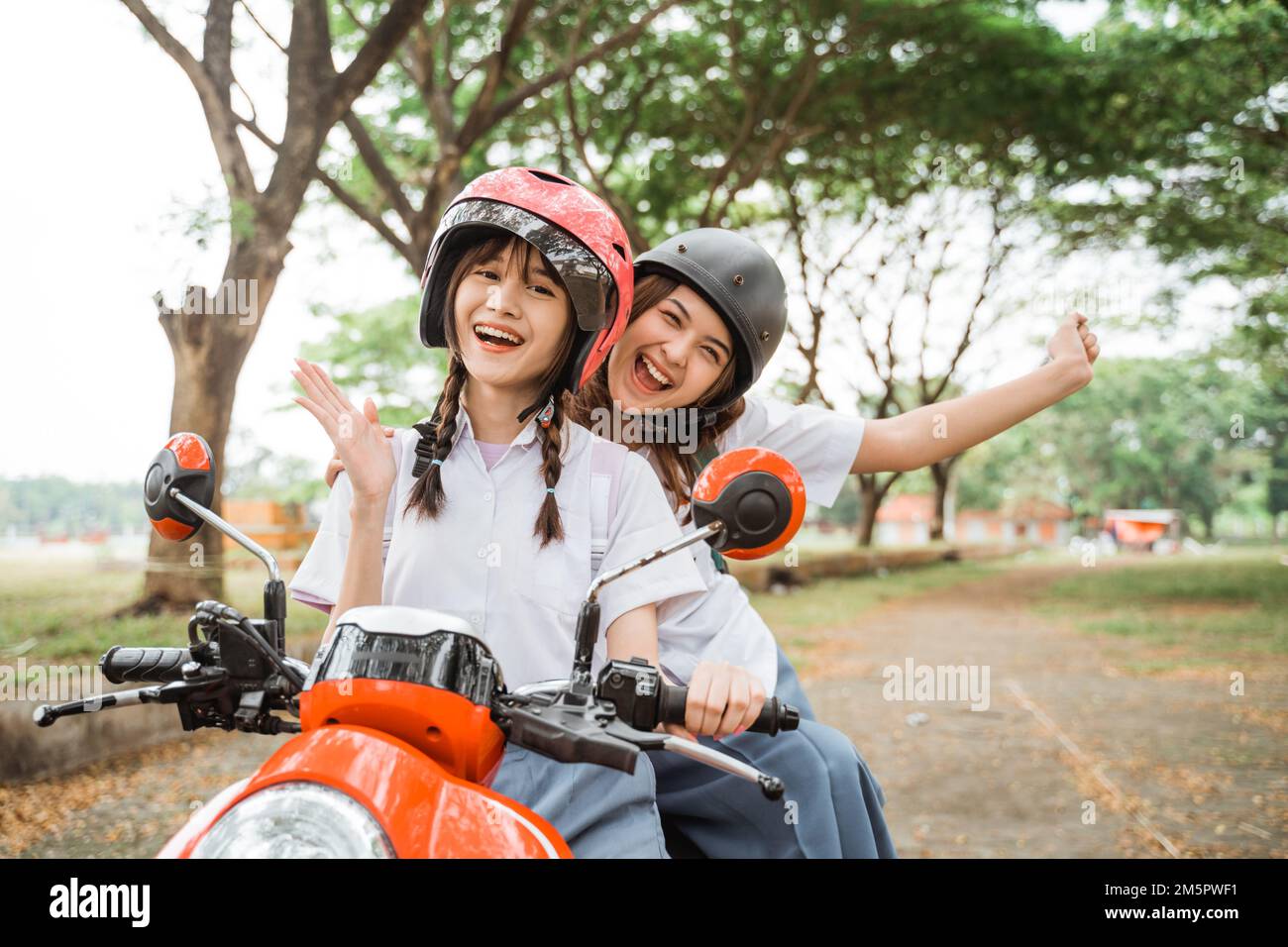 Two student girls wearing helmets waving while riding motorbikes Stock Photo