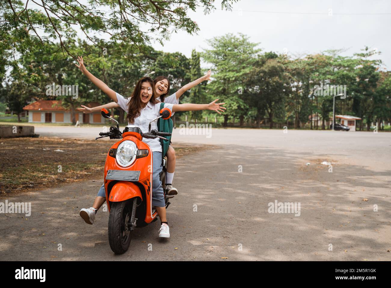 Two happy high school girls riding motorbikes waving hands Stock Photo