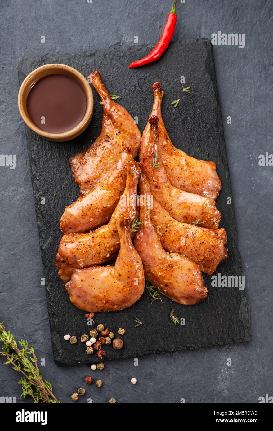 Delicacies. Grilled BBQ quail legs in marinade and spices on black slate with fresh herbs and sauce on a dark background. Healthy diet food. Top view. Stock Photo