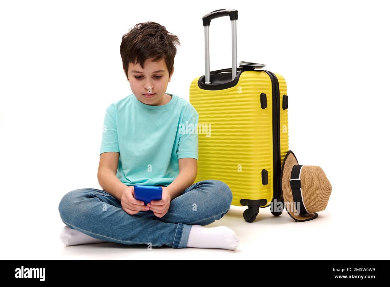 Teenager boy in blue t-shirt, with yellow suitcase, scrolling news feed, checking social media content on his smartphone Stock Photo