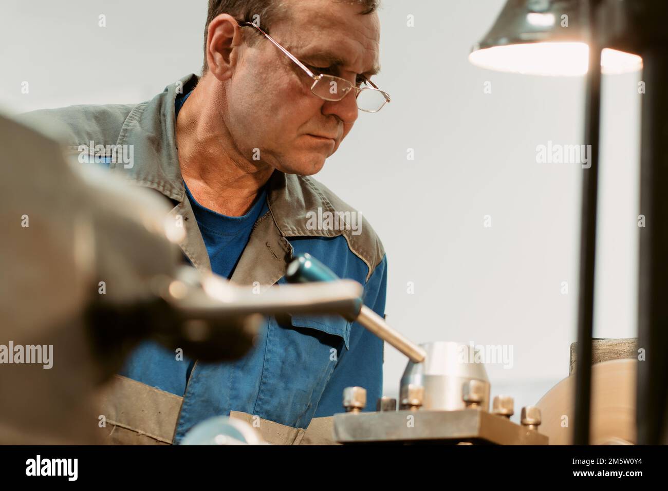 Portrait of turner with glasses 50-55 years old at work. Elderly metal turner works in workshop on machine behind job.. Stock Photo