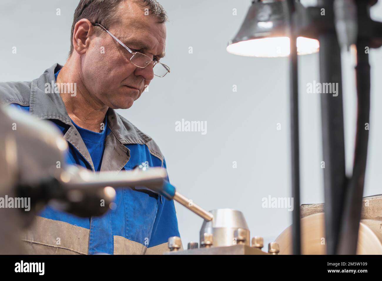 Portrait of turner with glasses 50-55 years old at work. Elderly metal turner works in workshop on machine behind job.. Stock Photo