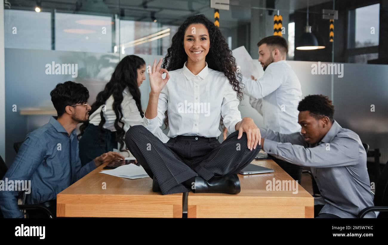 Happy calm smiling businesswoman female leader woman sitting on table in lotus position meditating in office showing ok hand gesture okay sign on Stock Photo