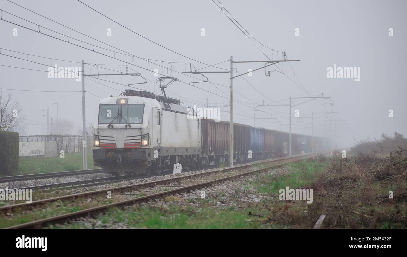 Freight train with a modern electric locomotive pulling open goods wagons or gondolas through foggy weather. Stock Photo