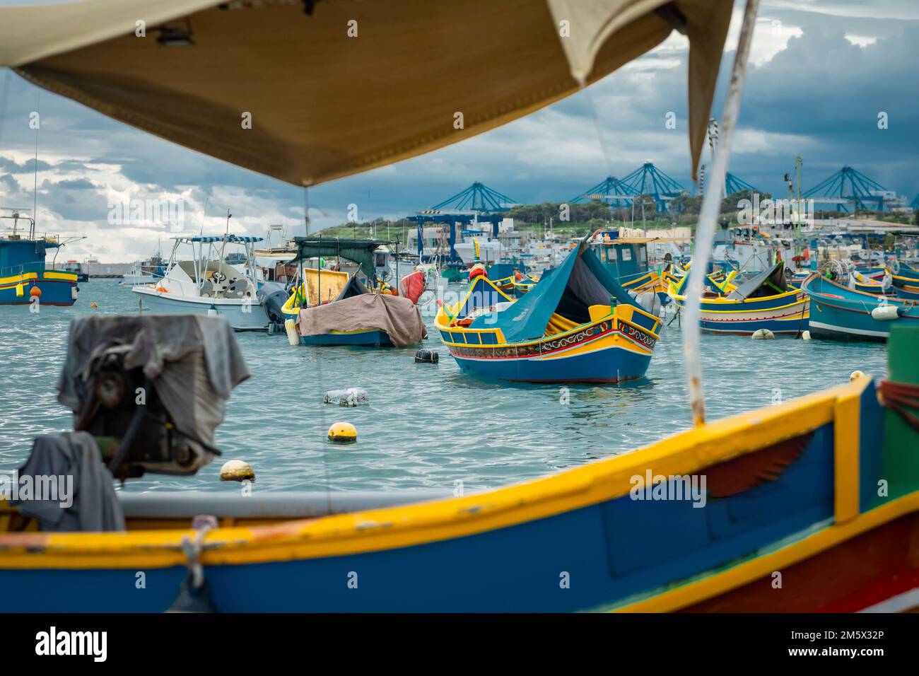 Luzzu, typical fishing boats of Malta moored in the marina of Marsaxlokk on an autumn day. Many ships in the bay, container cranes in the background. Stock Photo
