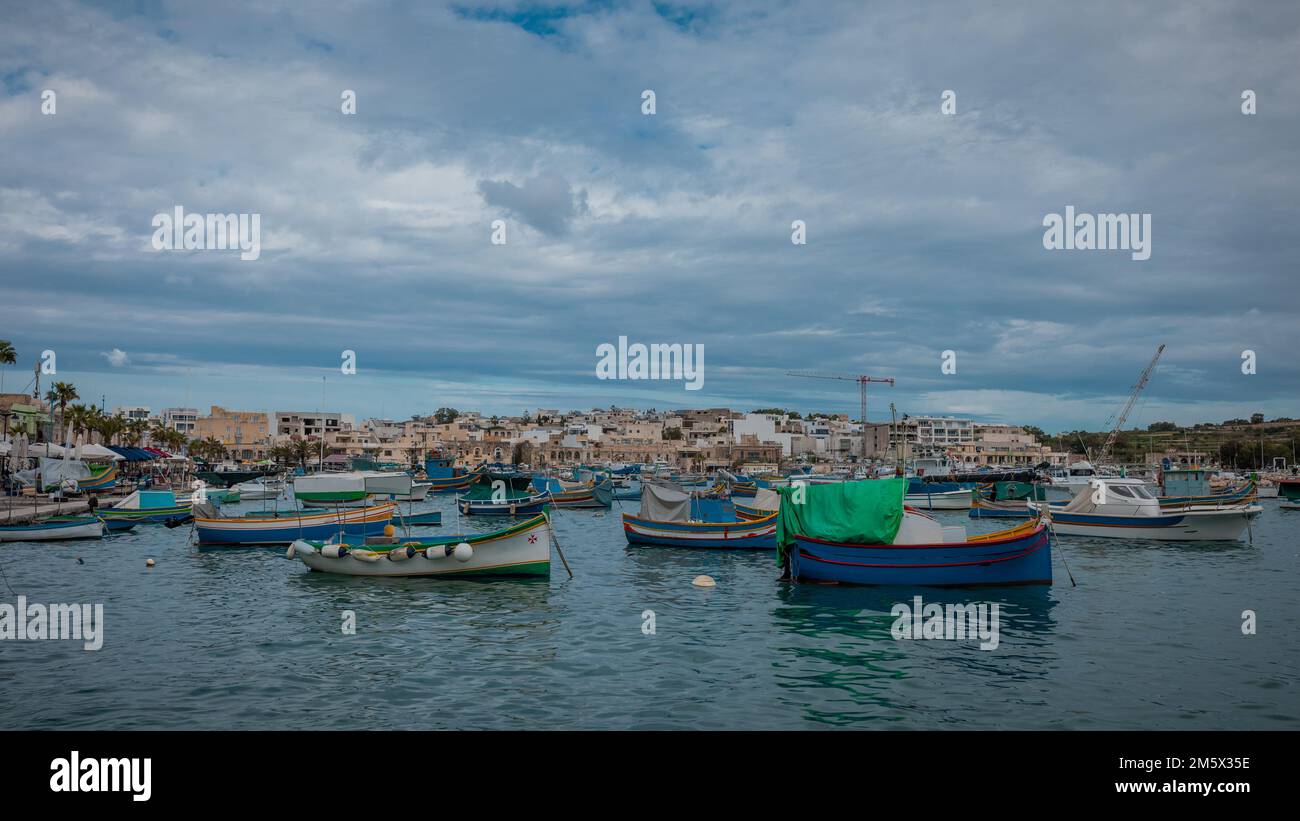 Luzzu, typical fishing boats of Malta moored in the marina of Marsaxlokk on an autumn day. Many ships in the bay, cityscape in the background. Stock Photo