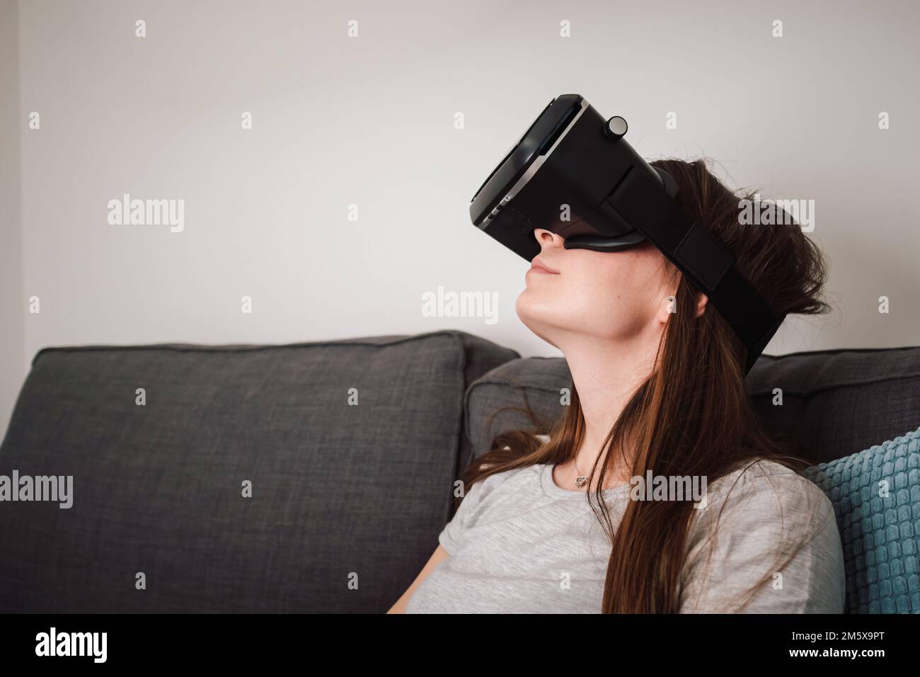 Beautiful smiling young woman wearing VR goggles and virtually traveling through the metaverse from the comfort of her couch Stock Photo