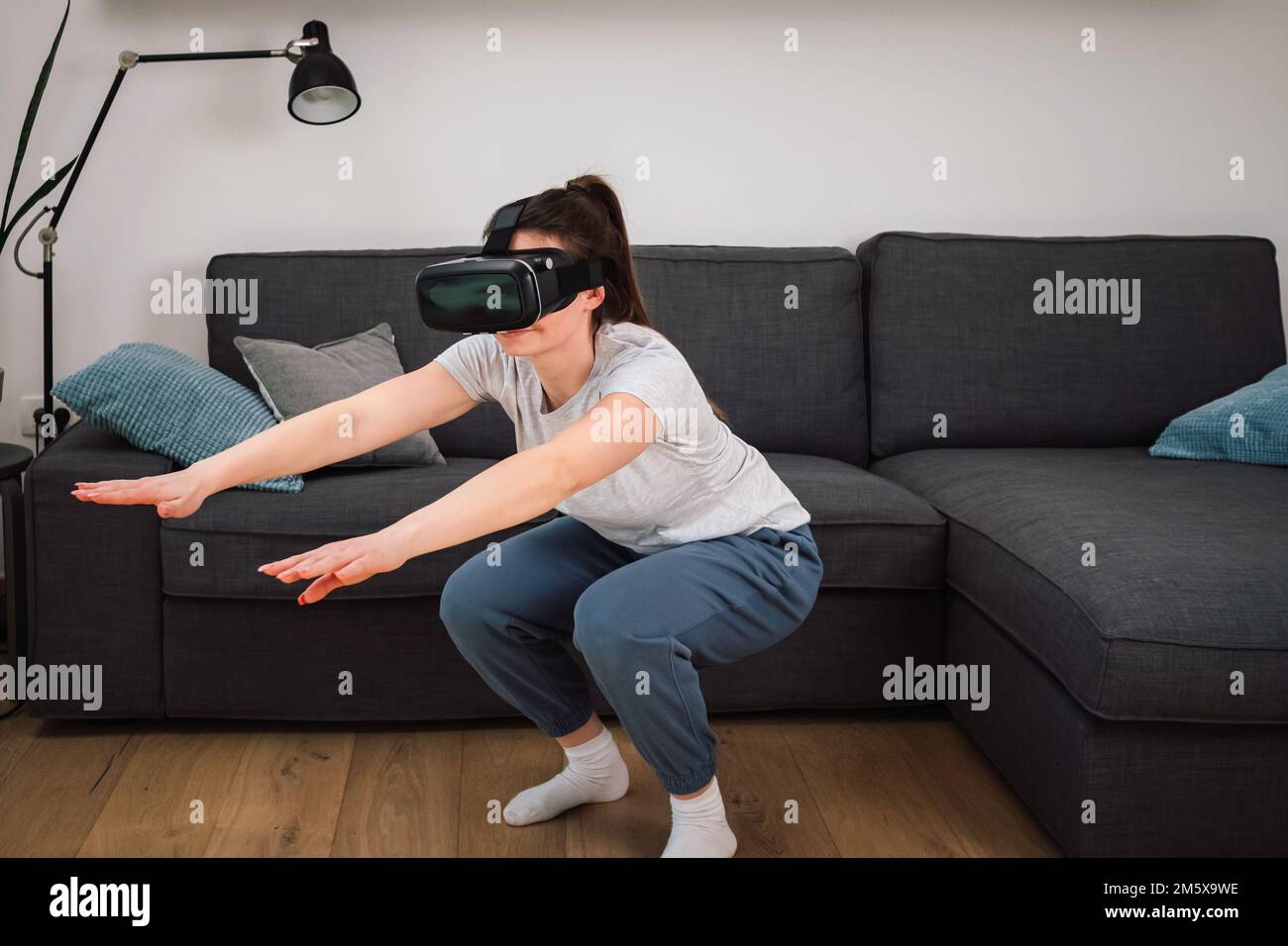 Caucasian young woman having a workout in VR headset, doing arm stretching exercises at home. Sport, health, and virtual reality concept. Stock Photo