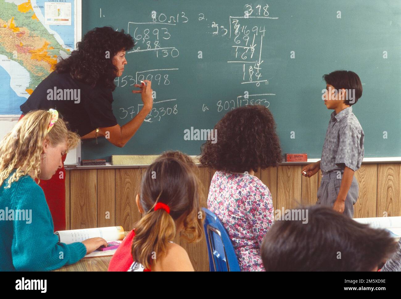 Teacher at the blackboard, writing, math problems. Well, a boy student stands trying to figure out the answers Stock Photo