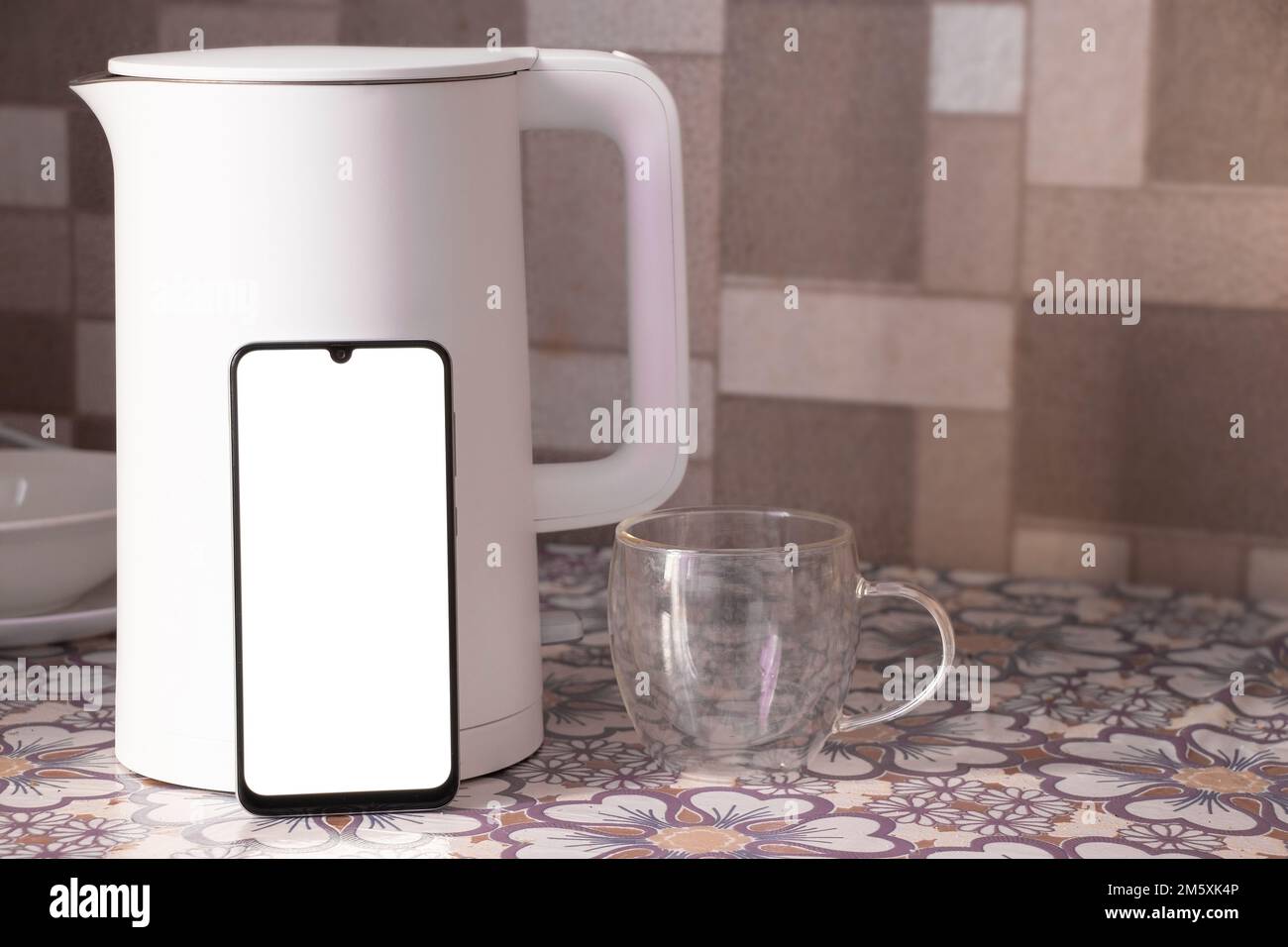 a phone with a blank white screen stands near a white teapot and a cup on the kitchen table Stock Photo