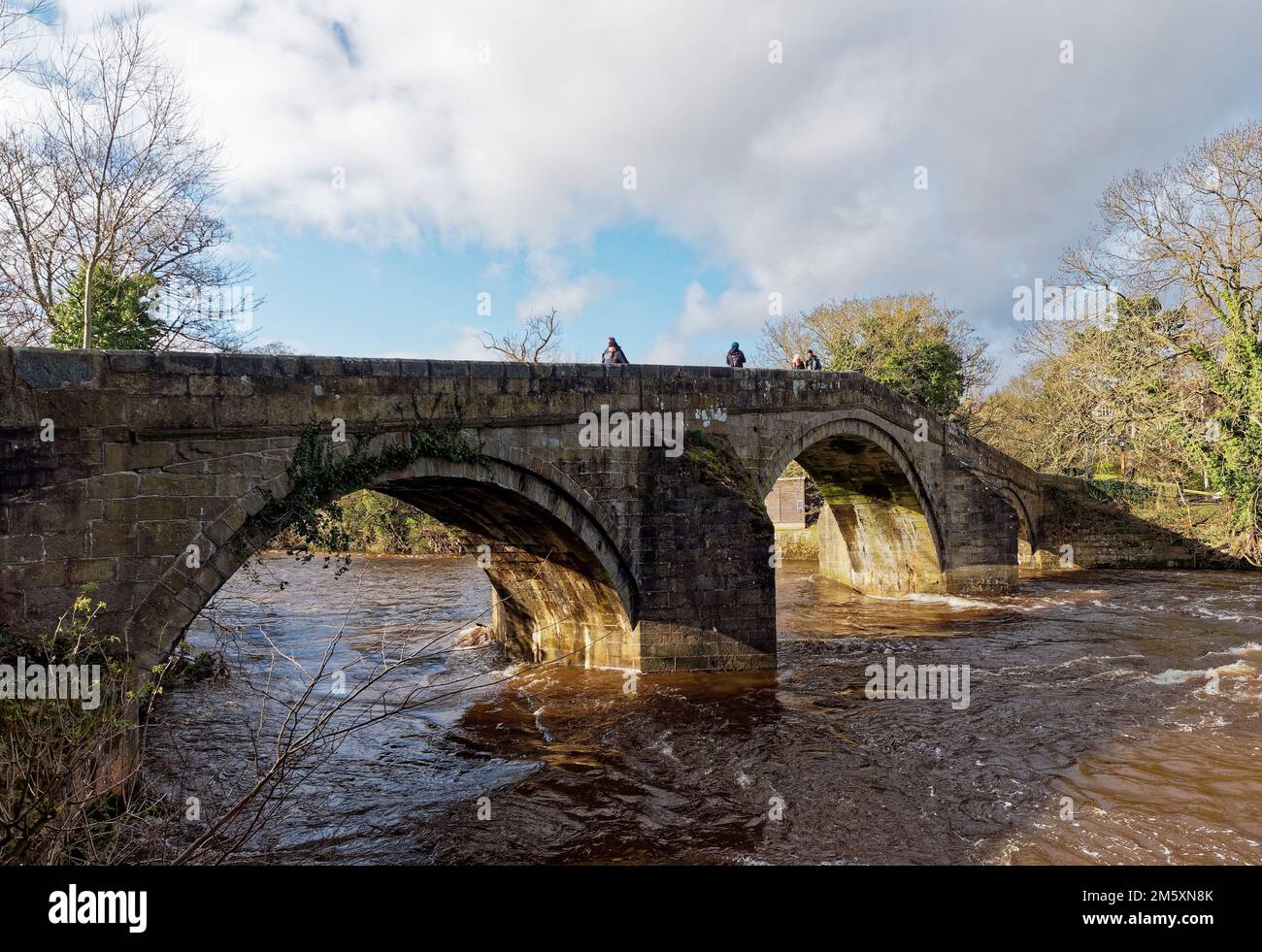 The old Stone Footbridge crowing the River Wharfe at Ilkley in West Yorkshire on a sunny cold day in March, with Walkers crossing it on the Dales way. Stock Photo
