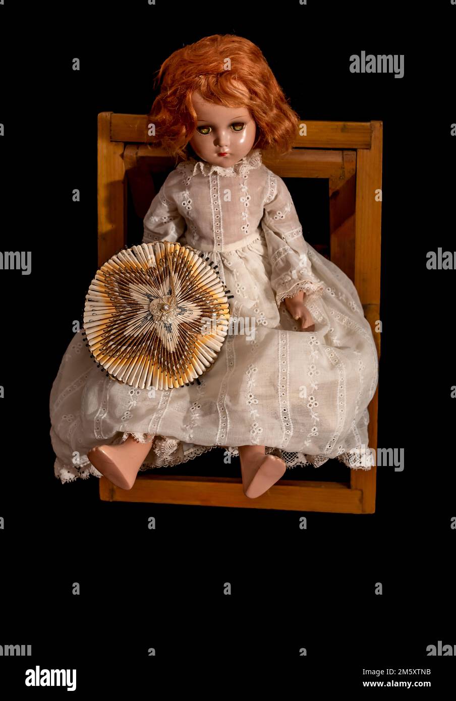 A vintage child’s doll, sitting in a wooden chair, with red hair, wearing a white dress and a holding vintage umbrella made from cigarette packages. Stock Photo