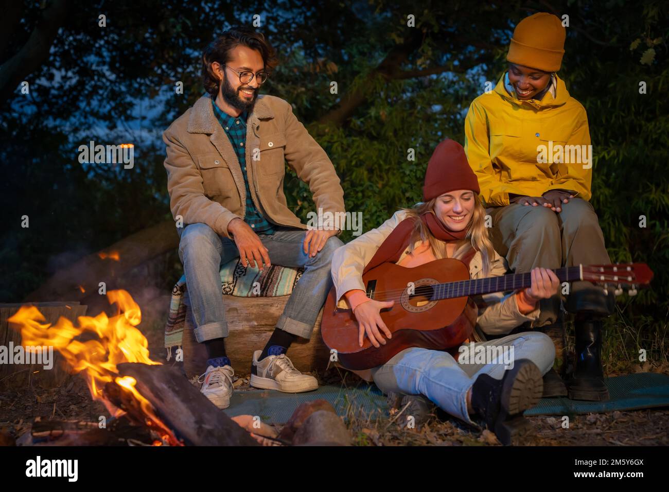 Three happy diverse friends having fun playing music and enjoying bonfire camping in nature at night Stock Photo