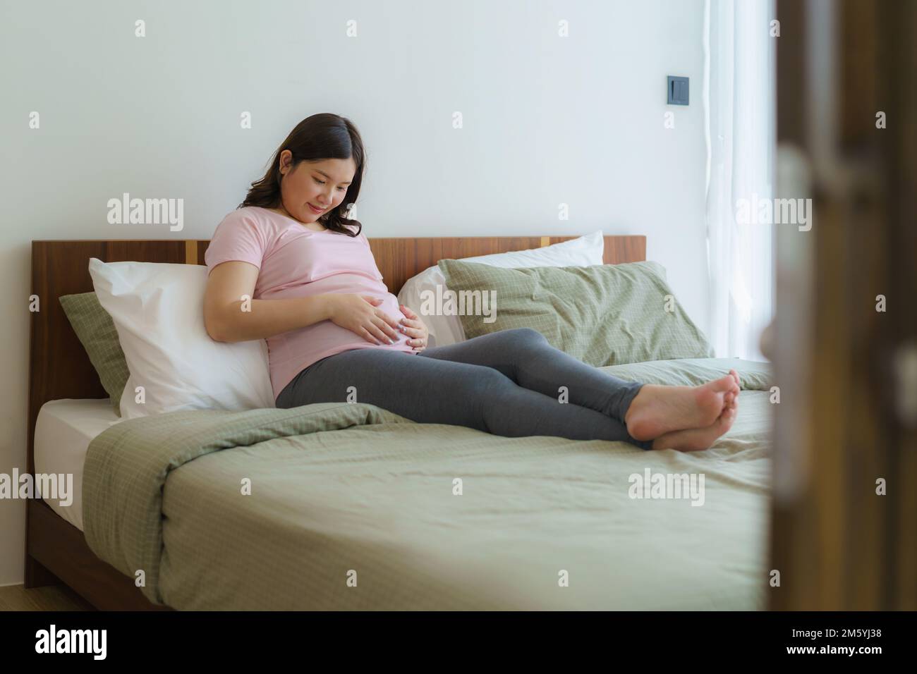 Pregnancy, people rest and expectation concept - Asian smiling happy pregnant woman sitting in bed and touching her belly at home. Stock Photo