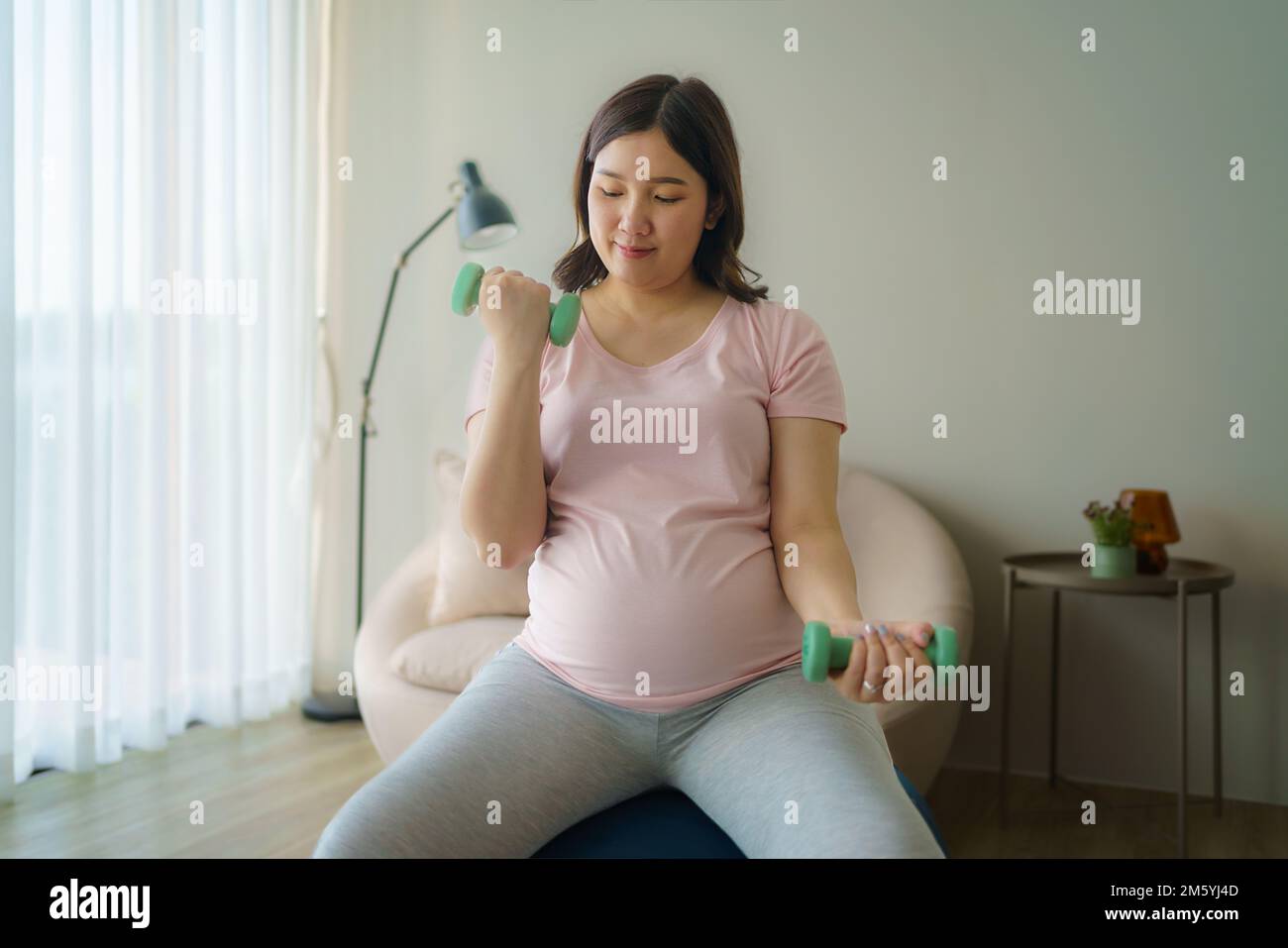 Beautiful Asian pregnant woman doing exercising with dumbbells in living room at home, healthy concept Stock Photo
