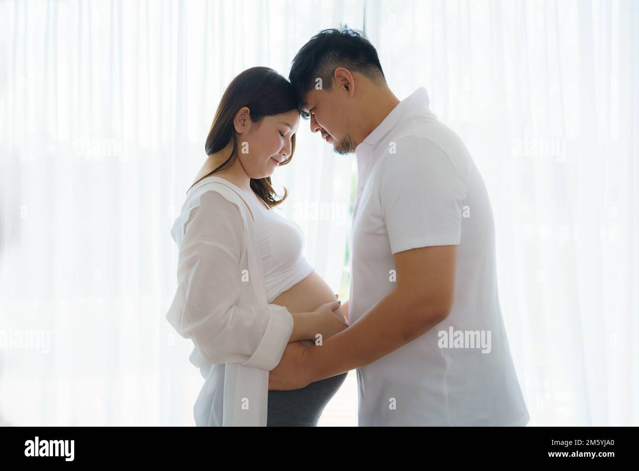 Happy Asian couple expecting baby standing together against window at home, loving husband tenderly touching belly of his pregnant wife Stock Photo