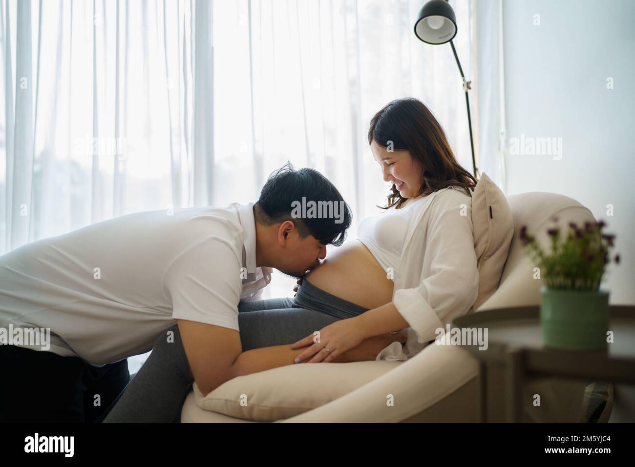 Happy Asian pregnant woman with husband expecting baby at home and husband try to kiss the baby through belly Stock Photo