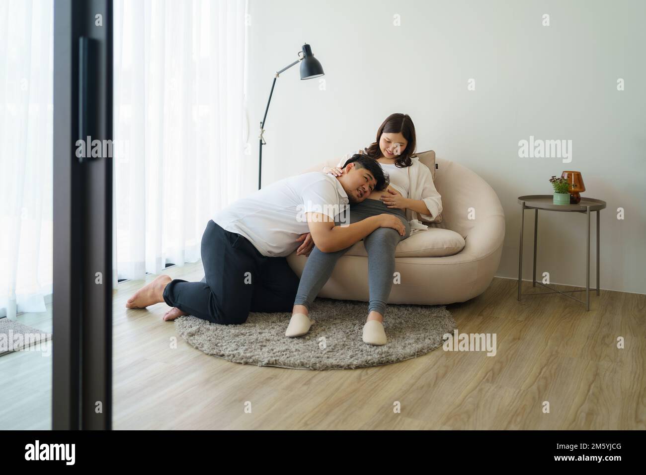 Happy Asian pregnant woman with husband expecting baby at home and husband try to kiss the baby through belly Stock Photo