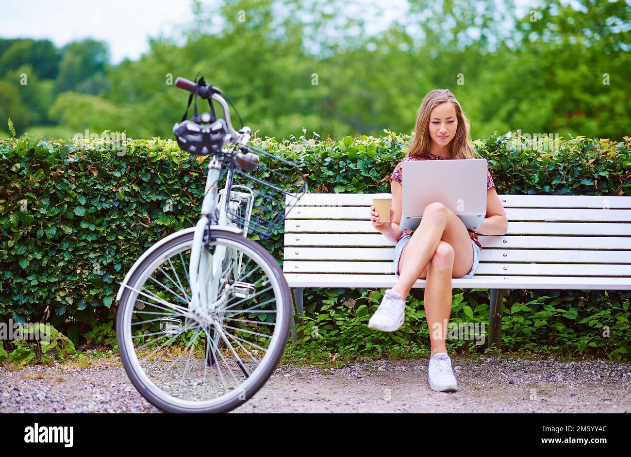 Blogging in the park. a young woman using a laptop on a bench while out for a cycle in the park. Stock Photo