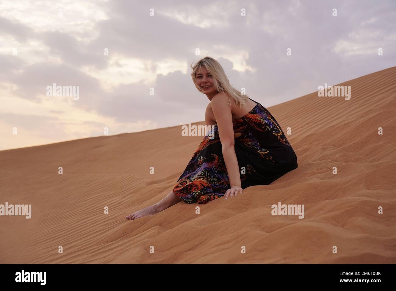 Beautiful blonde woman in a black dress is sitting on the hot sand in the desert. Beauty and travel concept. She smiles and looks at the camera Stock Photo