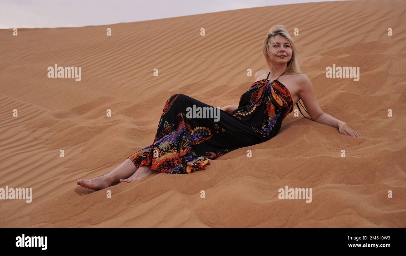 A young blonde lies on a pesce in the desert. Woman in the dunes. Travel and beauty. Wind in her hair Stock Photo